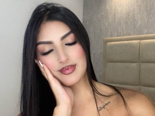 AriiaMon with long black hair, closed eyes, and pink lipstick, resting her hand on her cheek, showing a small tattoo on her neck, and her tits are not visible.