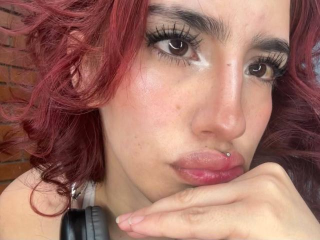 Elodie Roussee tiene el pelo rojo y rizado, un piercing en la nariz y está haciendo una mueca con la mano cerca de los labios. Lleva auriculares negros. Sus ojos están medio abiertos..