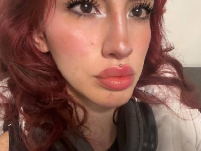 Elodie Roussee ha i capelli rossi e ricci, un piercing al naso e labbra rosa lucide. Indossa delle cuffie nere con una camicia bianca, e ha delle lentiggini evidenti e un trucco per gli occhi..