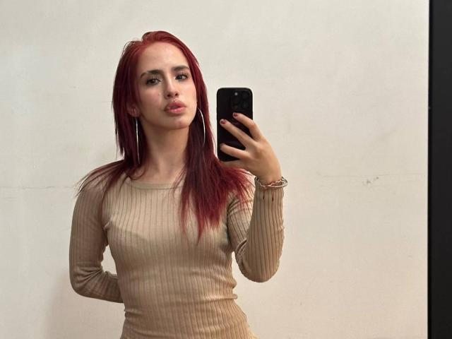 Elodie Roussee, avec des cheveux roux, portant un haut beige côtelé à manches longues, tenant un téléphone noir, faisant légèrement la moue, portant des boucles d'oreilles cerceaux et un bracelet, debout devant un mur uni..