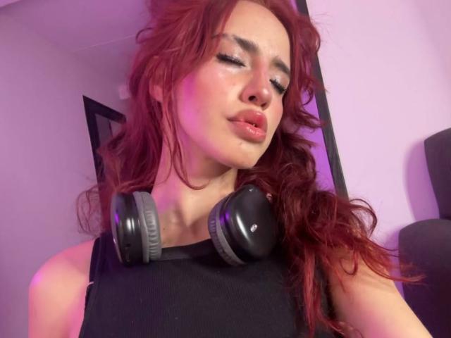 Elodie Roussee, con cabello rojo y ondulado, un top negro y auriculares negros, tiene una piel brillante y labios carnosos, mirando ligeramente hacia un lado con una expresión relajada..