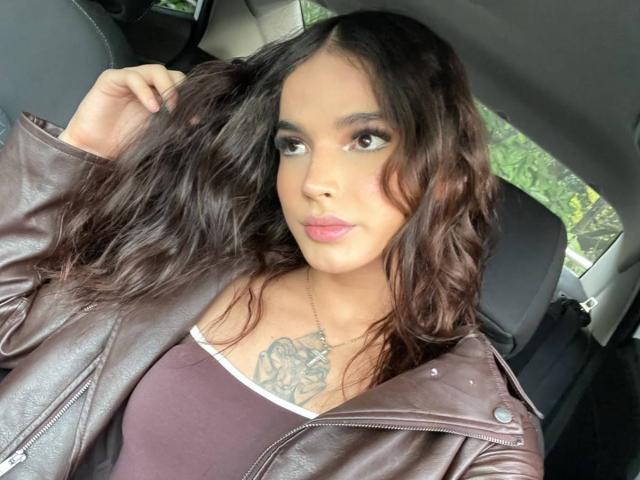 KateBmoss, con cabello castaño ondulado, vistiendo una chaqueta de cuero marrón y una camiseta sin mangas rosa, mostrando un tatuaje en el pecho, sentada en un coche, mirando a la cámara con labios rosados..