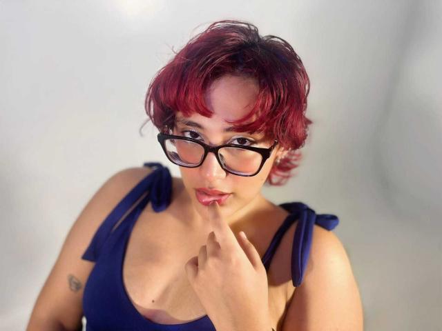 ChloeSterling with red-dyed short hair, black glasses, and a blue dress, touching her lips with her finger, showing a small tattoo on her upper arm.