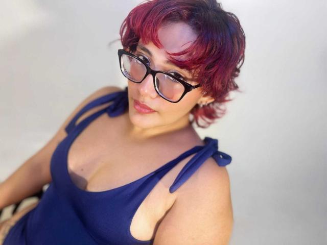 ChloeSterling with short red-purple hair, black glasses, and a blue dress with side ties, showing her tits and smiling slightly. She has light skin and pink lipstick.