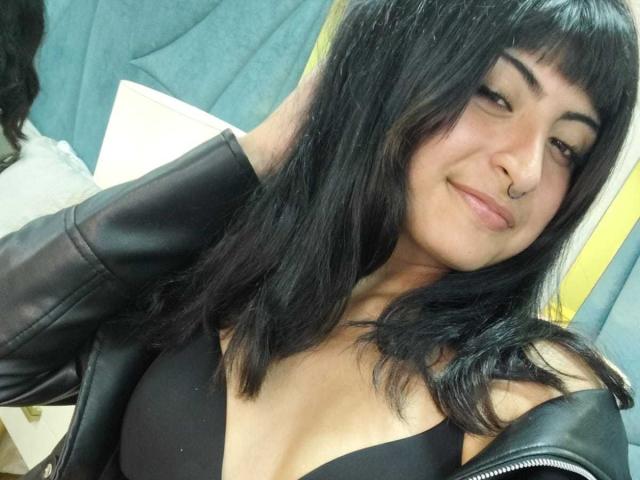MiaSalim with black hair, nose ring, and black leather jacket, smiling at camera, showing cleavage in black top, hands in hair. Boobs visible, slightly parted lips.