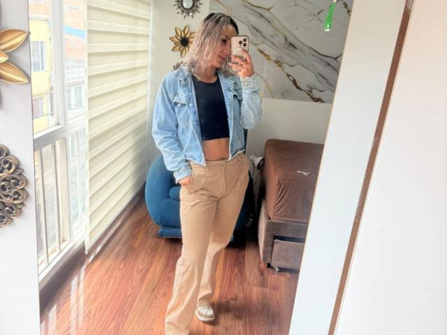 ElenaSaenz in a mirror selfie, wearing a blue denim jacket, black crop top, and tan high-waisted pants, with hands in pockets, blonde hair, wooden floor, and a brown couch.