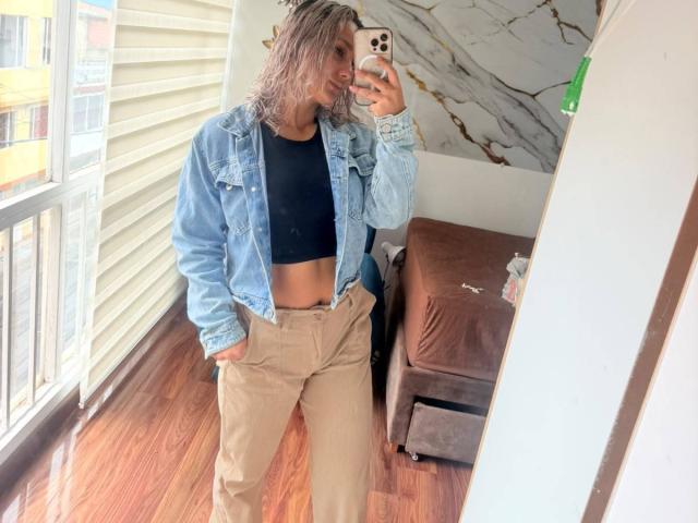 ElenaSaenz with wet blonde hair, denim jacket, black crop top, tan pants, and hands in pockets, taking a mirror selfie, showing a toned, flat stomach.