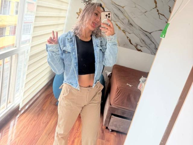 ElenaSaenz with wavy blonde hair, denim jacket, black crop top, and tan pants, making a peace sign, tongue out, in a mirror selfie, standing on wooden floor.