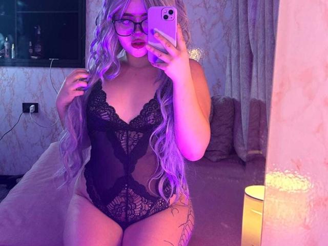 Lyanekkoo, con cabello largo morado, gafas y lencería de encaje negro, mostrando sus grandes senos y su trasero curvilíneo, tomando un selfie frente al espejo con iluminación rosa y morada..