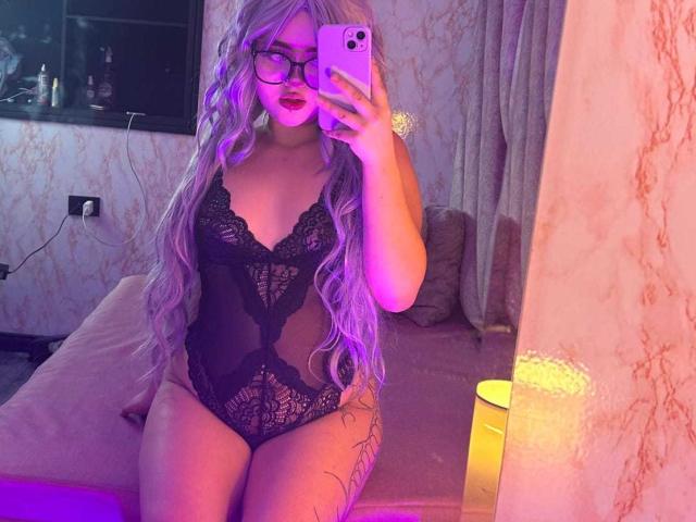 Lyanekkoo with long purple hair, glasses, and black lace lingerie, showing her pussy through the lace, sitting on a bed, taking a mirror selfie.