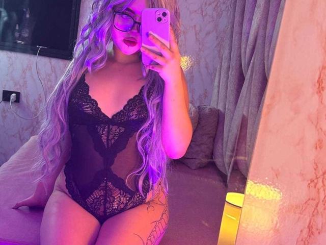Lyanekkoo with purple hair, glasses, and black lace bodysuit, taking a mirror selfie showing her tits and tattooed thigh, in pink and purple lighting.