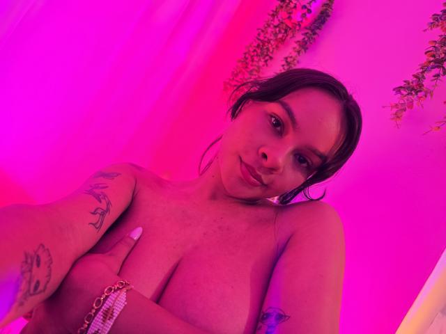Yurimy with pink-tinted skin, dark hair, and tattoos, covering her small tits with a hand, wearing a bracelet, looking at the camera with a slight smile.