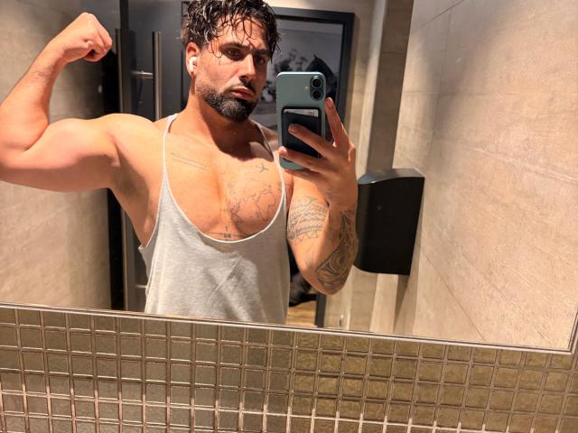 Muscular Gasston69 with wet, dark hair flexing bicep, wearing a grey tank top, showing tattoos, holding a phone in a mirror selfie, showing his chest and arm.