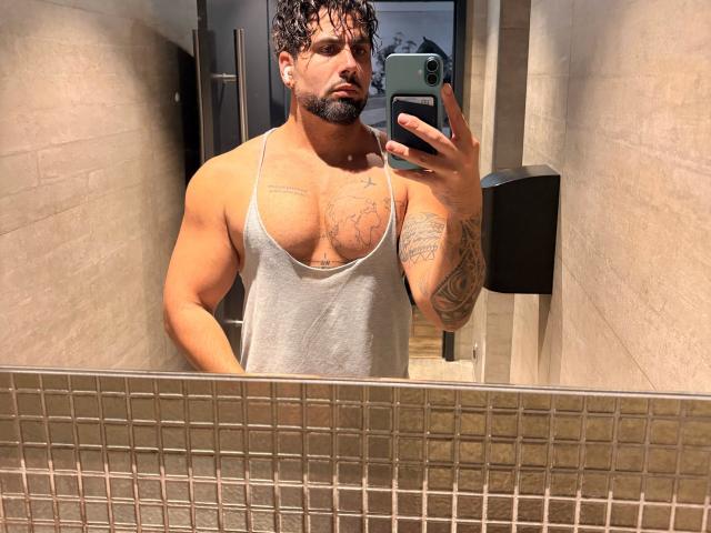 Muscular Gasston69 with curly hair and beard, wearing a grey tank top, showing tattoos on chest and arm, taking a mirror selfie in a bathroom with tiled mirror.