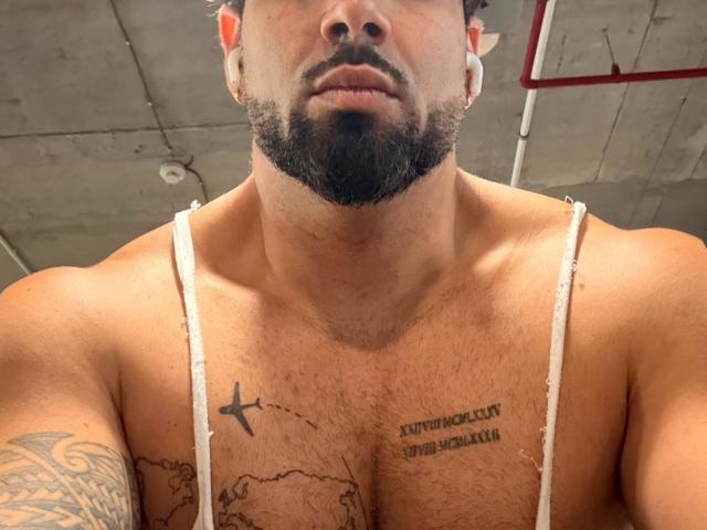 Gasston69's muscular chest with tattoos, wearing a white tank top, has a cross tattoo on the left and a map tattoo on the right, with a beard and short hair.
