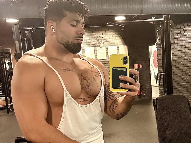Muscular Gasston69 with tan skin, black beard, white tank top showing big tits, tattoos on chest and arm, holding yellow phone, taking mirror selfie in gym.