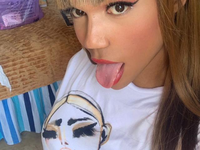 KendallKiss has tan skin, long blonde hair, and is sticking out her tongue with red lipstick. She's wearing a white shirt with a black-and-white portrait graphic.