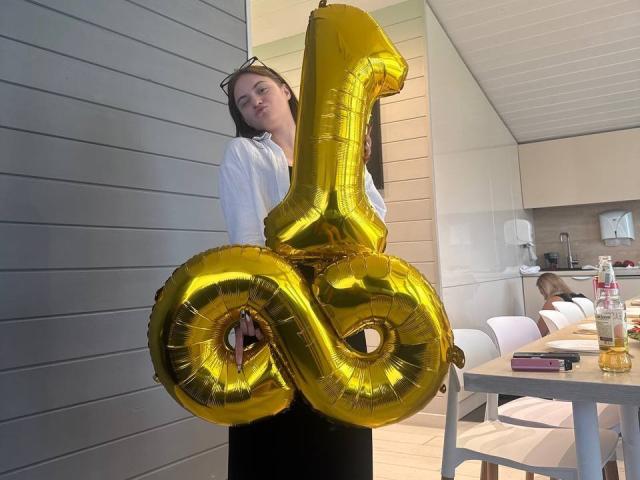 FriedaArnaldo holding shiny golden number 8 balloons, wearing white shirt and black skirt, standing in modern kitchen with white chairs and table, beer bottle on right.