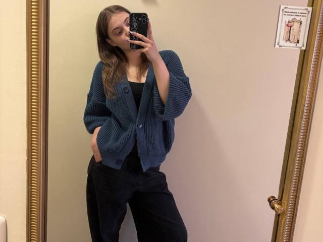 FriedaArnaldo with medium tits, wearing a blue knit cardigan, black top, and black pants, hands in pockets, taking a mirror selfie, brown hair, gold-framed mirror.