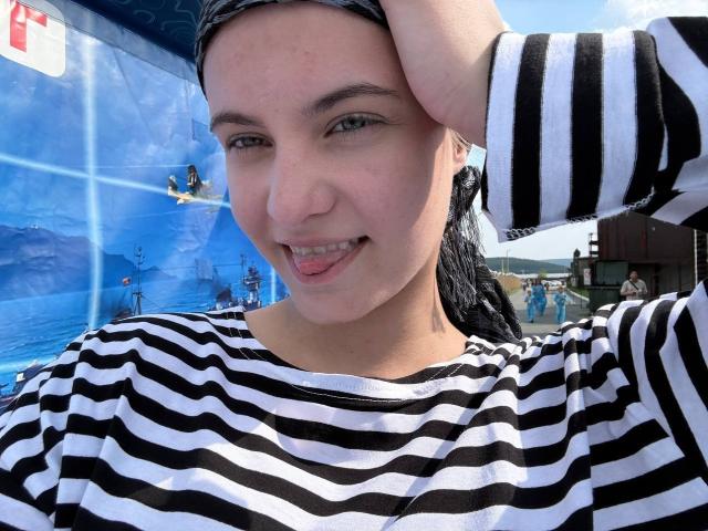 FriedaArnaldo with black and white striped shirt, hand in hair, tongue out, smiling, blue eyes, fair skin, black headscarf, water background, looking cute, showing teeth.