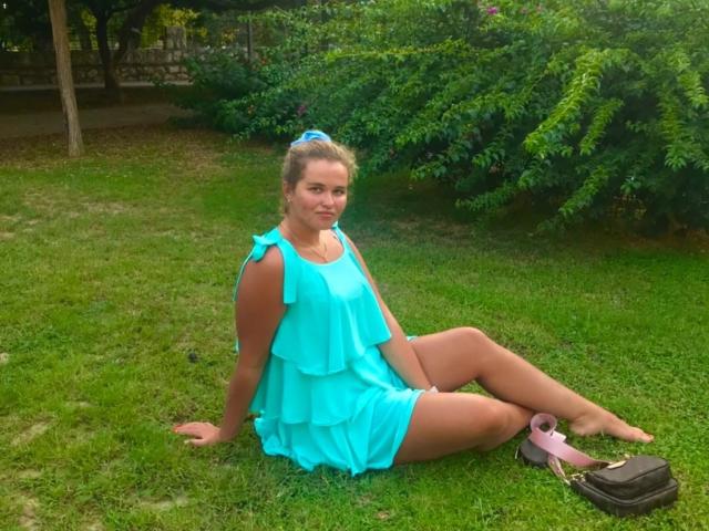 JessicaJoye is sitting on grass, wearing a turquoise ruffled dress, her bare ass exposed, blonde hair in a ponytail, pink shoes on the ground, green bush behind her.