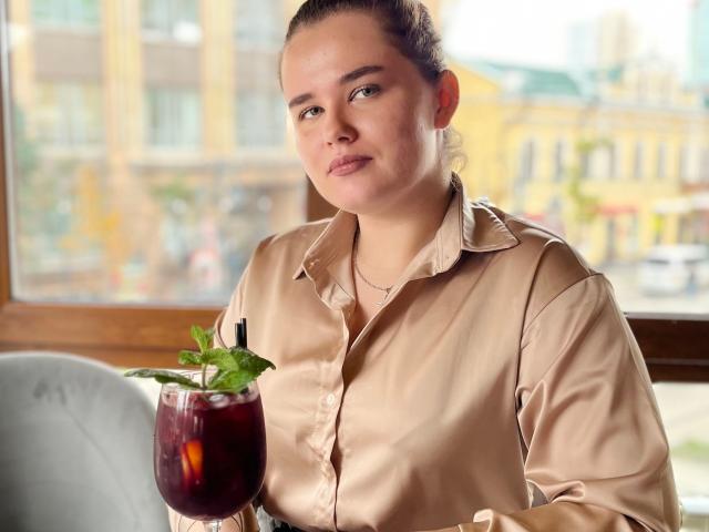 JessicaJoye with light brown shirt, holding dark red drink with green mint, relaxed, slight smile, brown hair in bun, light skin, small necklace, indoors, sunlight through window.