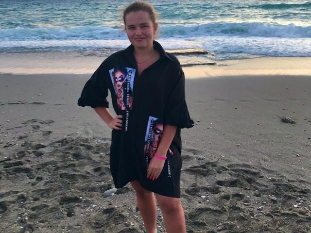 JessicaJoye standing on a beach, wearing a black dress with colorful prints, hands on hips, blonde hair in a messy bun, pink wristband, ocean and waves in the background.