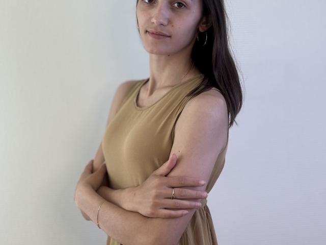 RobbynRoseum, mit einem beigen Kleid, Armen verschränkt, braunen Haaren, einem leichten Lächeln, kleinen Brüsten, schlanker Figur, Reifenohrringen, einem Ring, heller Haut, stehend, weißer Hintergrund, weiches Licht, lässige Pose..