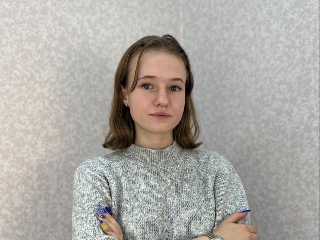 MadgeShowell with light brown hair, green eyes, and grey knit sweater, arms crossed, blue nails, small earrings, and neutral expression, standing against a grey textured wall.