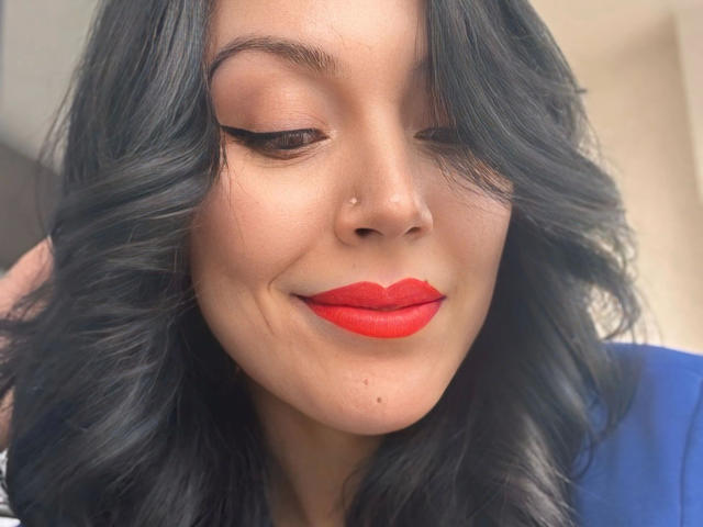 Layla84-hot has wavy black hair, red lipstick, and a small nose piercing. She's smiling with closed eyes, wearing black eyeliner and a blue top.