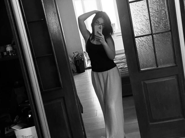 Black-and-white mirror selfie of AlexXas with arms up, wearing a black tank top and white pleated pants, showing her tits and ass, holding a phone in a doorway.