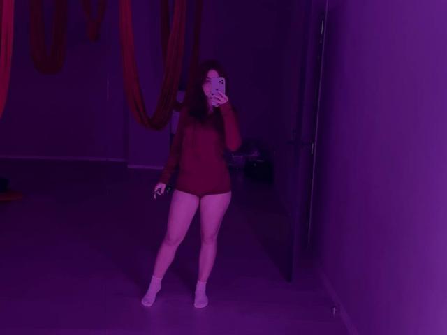 AlexXas with medium tits, wearing a red long-sleeve leotard, white socks, and no pants, taking a selfie in purple-lit room, covering her mouth with her phone.