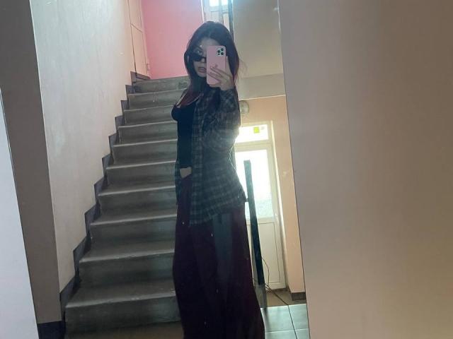 AlexXas, standing in a hallway, taking a mirror selfie, wearing a black top and long dark skirt, with hands in pockets, visible stairs behind, holding a pink phone.