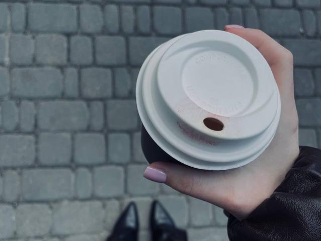 AlexXas's hand holding a white coffee cup lid with a brown hole, pale pink nails, black leather sleeve, black shoes partially visible.