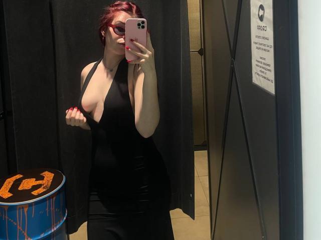 AlexXas with red hair, wearing a black, deep-cut dress, holding a phone in a mirror selfie, showing her tits, standing in a room with a blue trash can.
