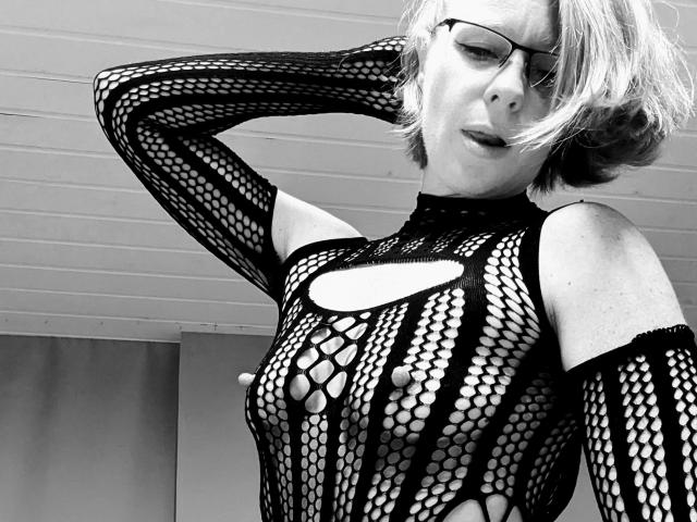 HeisseLina – eine attraktive Frau mit kurzen, blonden Haaren und Brille, die ein schwarzes Netzhemd trägt, durch das ihre Brüste sichtbar sind. Sie blickt nach unten und hat eine Hand in ihrem Haar..