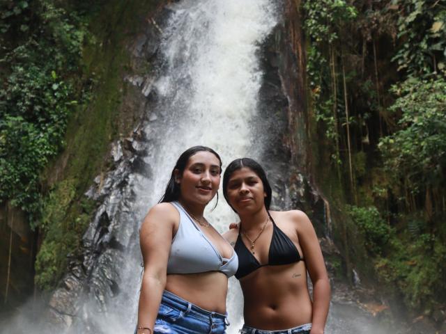 SaraAndGaby standing in front of a waterfall, both with wet hair, Sara in a light blue bra and jeans, Gaby in a black bra and jeans, showing their tits.