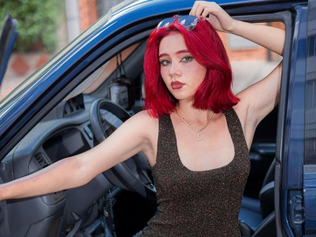 ChloeeWilliams with bright red hair, wearing a glittery black tank top, leaning on a car door with sunglasses on her head, showing her armpits and a gold necklace.