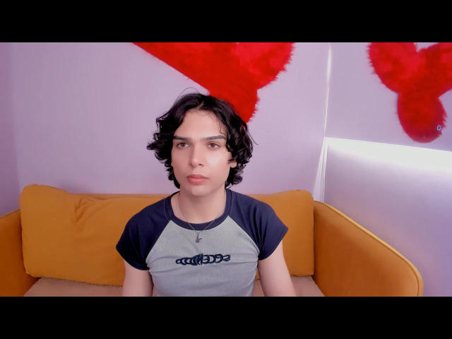 RubyMote with curly black hair, wearing a gray and black raglan shirt, sitting on an orange couch, has a neutral expression, and a small cross necklace.