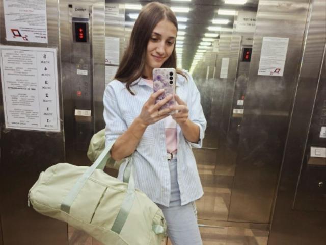 RobbynRoseum è in un ascensore di metallo, sta scattando un selfie con un telefono viola, indossa una camicia a righe blu, un reggiseno rosa e pantaloni bianchi, e tiene in mano una borsa verde..
