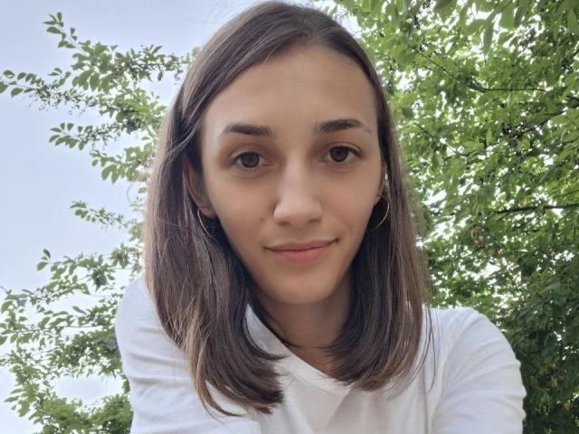 RobbynRoseum tiene el cabello castaño medio, ojos marrones y una ligera sonrisa, y lleva una camisa blanca, con pequeños aros, en un entorno exterior brillante con árboles de hojas verdes..