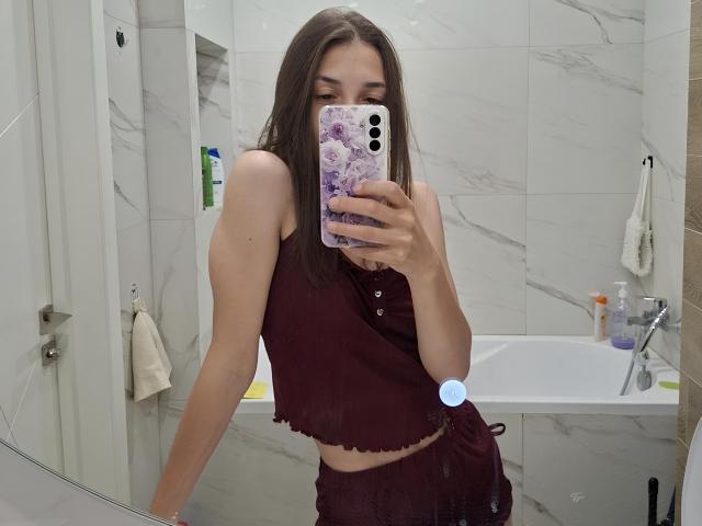 RobbynRoseum, avec de longs cheveux bruns, portant un débardeur bordeaux et un short assorti, prend un selfie devant un miroir avec une coque de téléphone violette, montrant une silhouette mince, plate et petite..