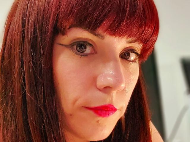 OrianaLaFrancaise with red lipstick, dark brown eyes, and winged eyeliner, has straight red hair with bangs, and a slight smirk on her face. Her nose is slightly shiny.