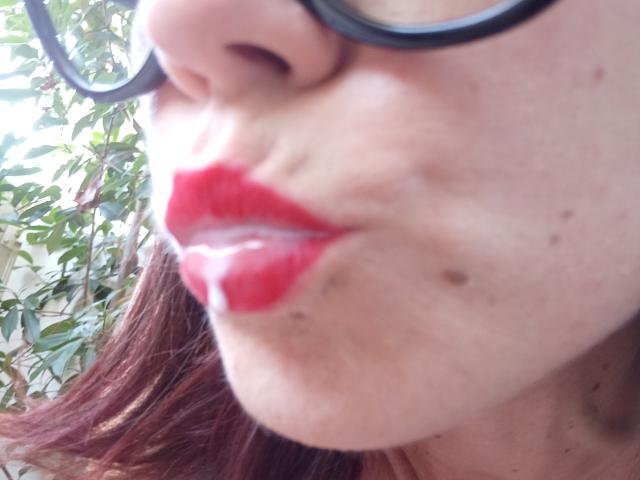 Het gezicht van OrianaLaFrancaise met rode lippenstift, een witte vloeistof op haar glanzende lippen, zwarte bril, licht open lippen, bruin haar en een paar kleine donkere moedervlekken op haar wang..