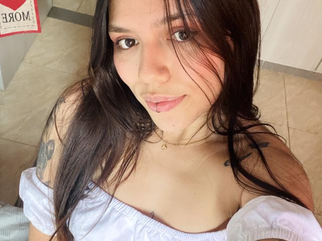 SophiaMarx with long brown hair, pink tongue out, wearing a white off-shoulder top, showing small tits and a tattoo on her right arm. She's looking at the camera.