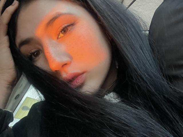 PattiTropea with long black hair, orange light on her right cheek, brown eyes, and slightly parted pink lips, wearing a black top, hand in hair.