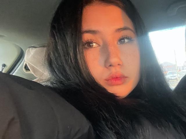 PattiTropea's face in a car, with long black hair, pouty pink lips, and light brown eyes, wearing a black shirt, sunlight on her right side.