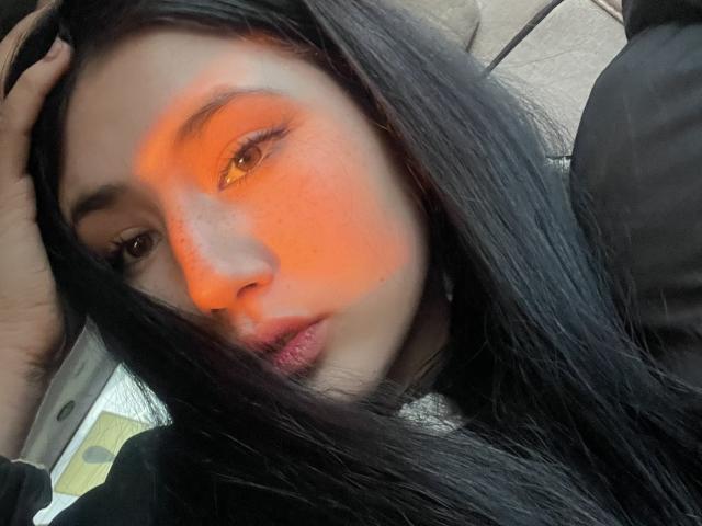PattiTropea has long black hair, orange glow on her face, brown eyes, and slightly parted lips. Her hand is in her hair. She's wearing a black top.
