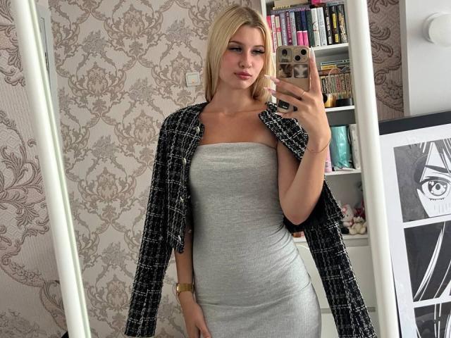 EmilyMorlino with blonde hair, gray strapless dress, black checkered jacket, holding phone, standing in mirror, showing her tits, in a stylish room.