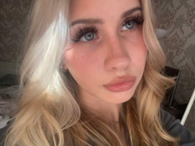EmilyMorlino has blonde hair, blue eyes, and full pink lips. She's wearing long, thick eyelashes and a gray shirt. Her face is slightly flushed with a cute, serious expression.